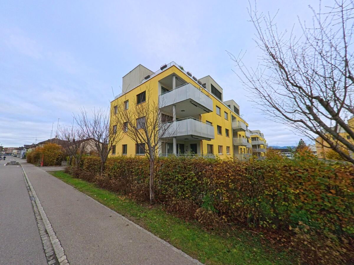 Mehrgeschossige gelbe Wohnbauten mit Balkonen stehen in Kirchberg (9602, Kanton St. Gallen) hinter einer herbstlichen Hecke entlang eines breiten Gehwegs.