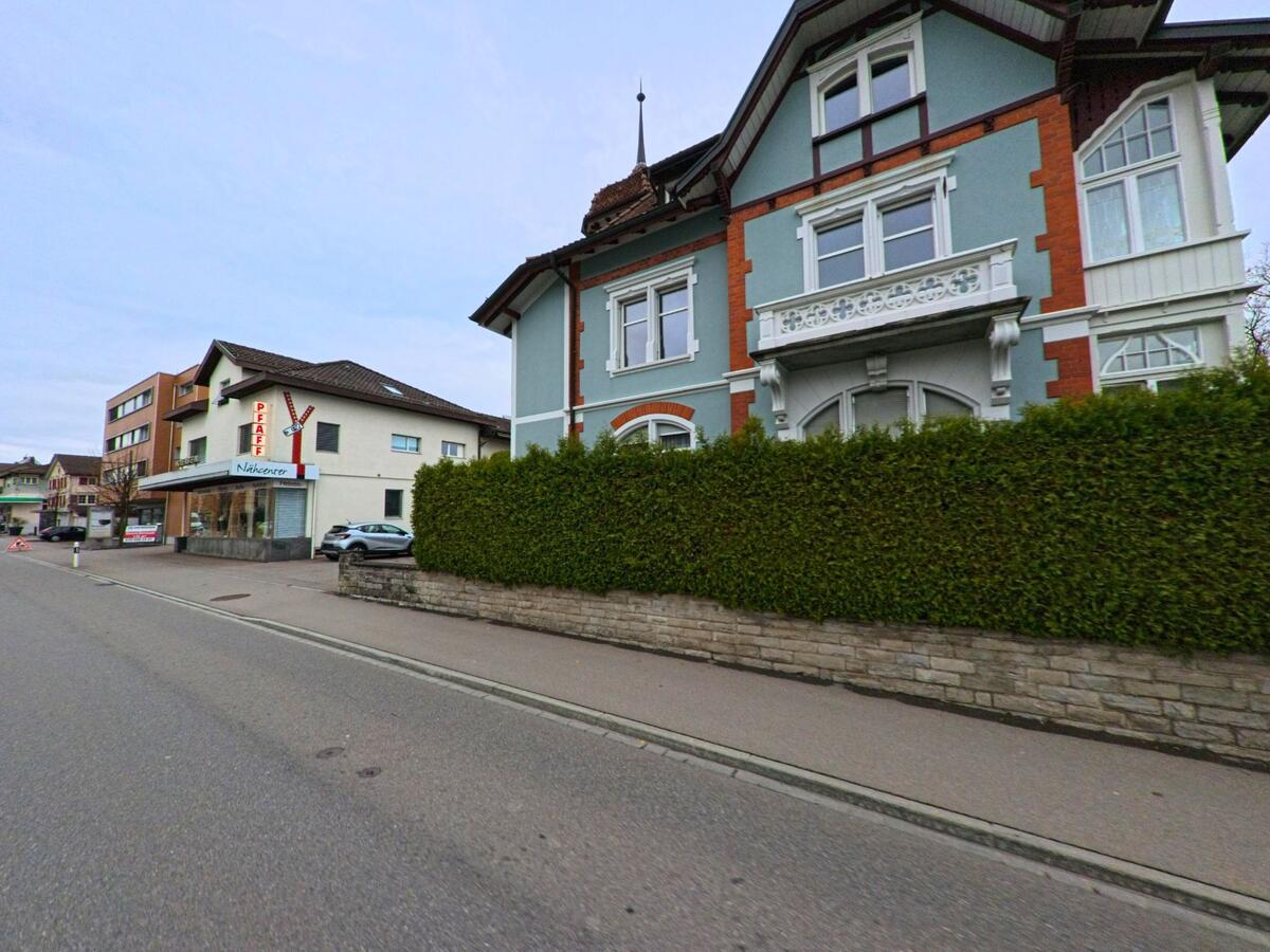 Ein hellblaues historisches Wohnhaus mit weissem Zierrat steht in Kirchberg (9602, Kanton St. Gallen) nahe einem kleinen Geschaeft an einer breiten Strasse.