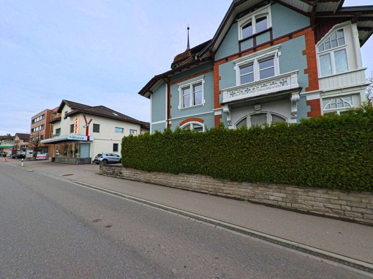Ein hellblaues historisches Wohnhaus mit weissem Zierrat steht in Kirchberg (9602, Kanton St. Gallen) nahe einem kleinen Geschaeft an einer breiten Strasse.
