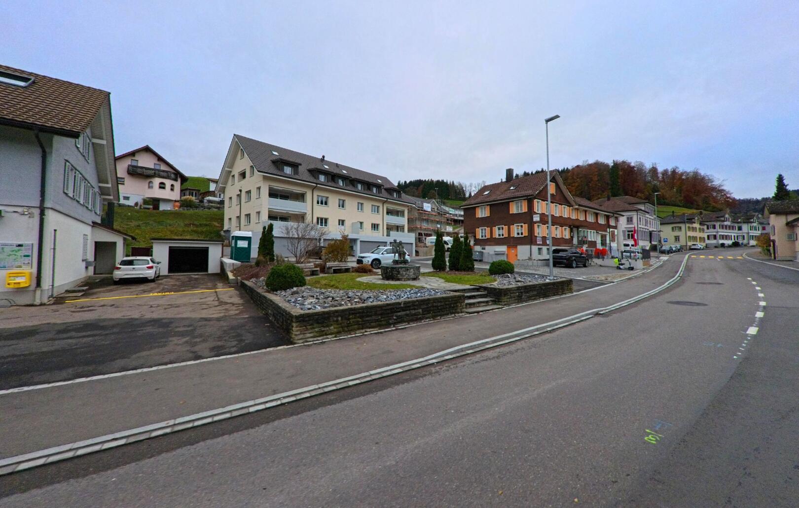 Zwischen Wohnhaeusern in Kirchberg (9602, Kanton St. Gallen) liegt ein kleiner Platz mit Skulptur und begruenten Beeten an einer sanft ansteigenden Strasse.