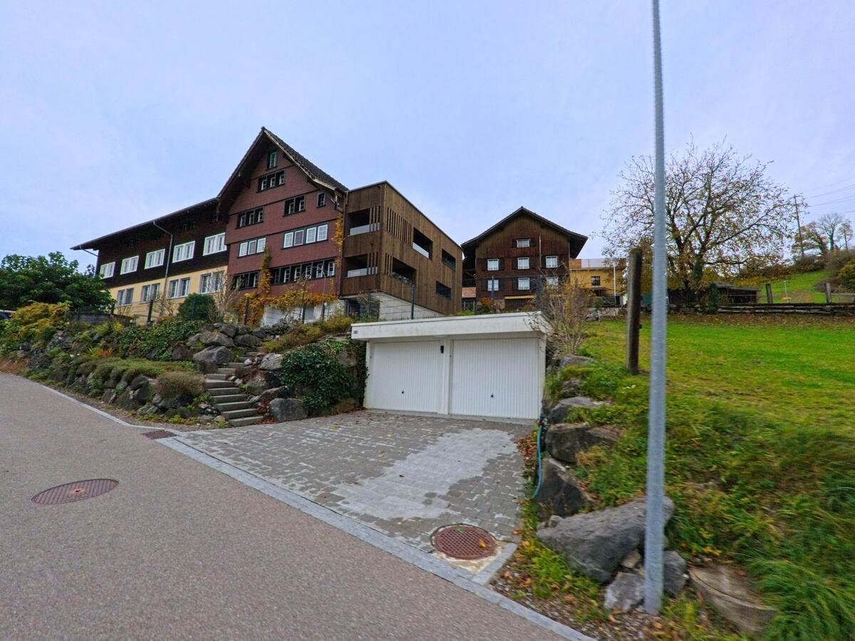 Mehrere traditionelle Holzhäuser stehen in Kirchberg (9602, Kanton St. Gallen) oberhalb einer Stützmauer mit Natursteinen, neben einer Garage und einer grossen Wiese.