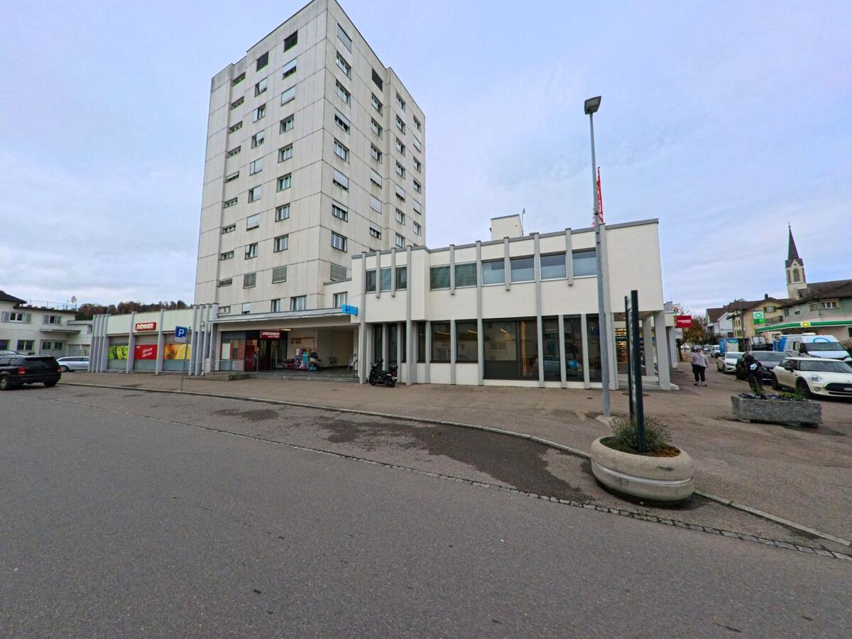 Ein hoher Wohn- und Geschaeftsbau praegt in Kirchberg (9602, Kanton St. Gallen) das Zentrum, wo Ladenfronten, Parkflaechen und Fussgaenger den Platz beleben.