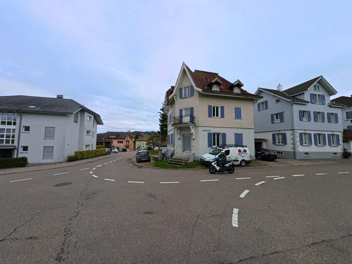 An einer breiten Kreuzung in Kirchberg (9602, Kanton St. Gallen) stehen helle Wohnhaeuser mit blauen Fensterlaeden, waehrend Autos und ein Motorrad vorbeifahren.