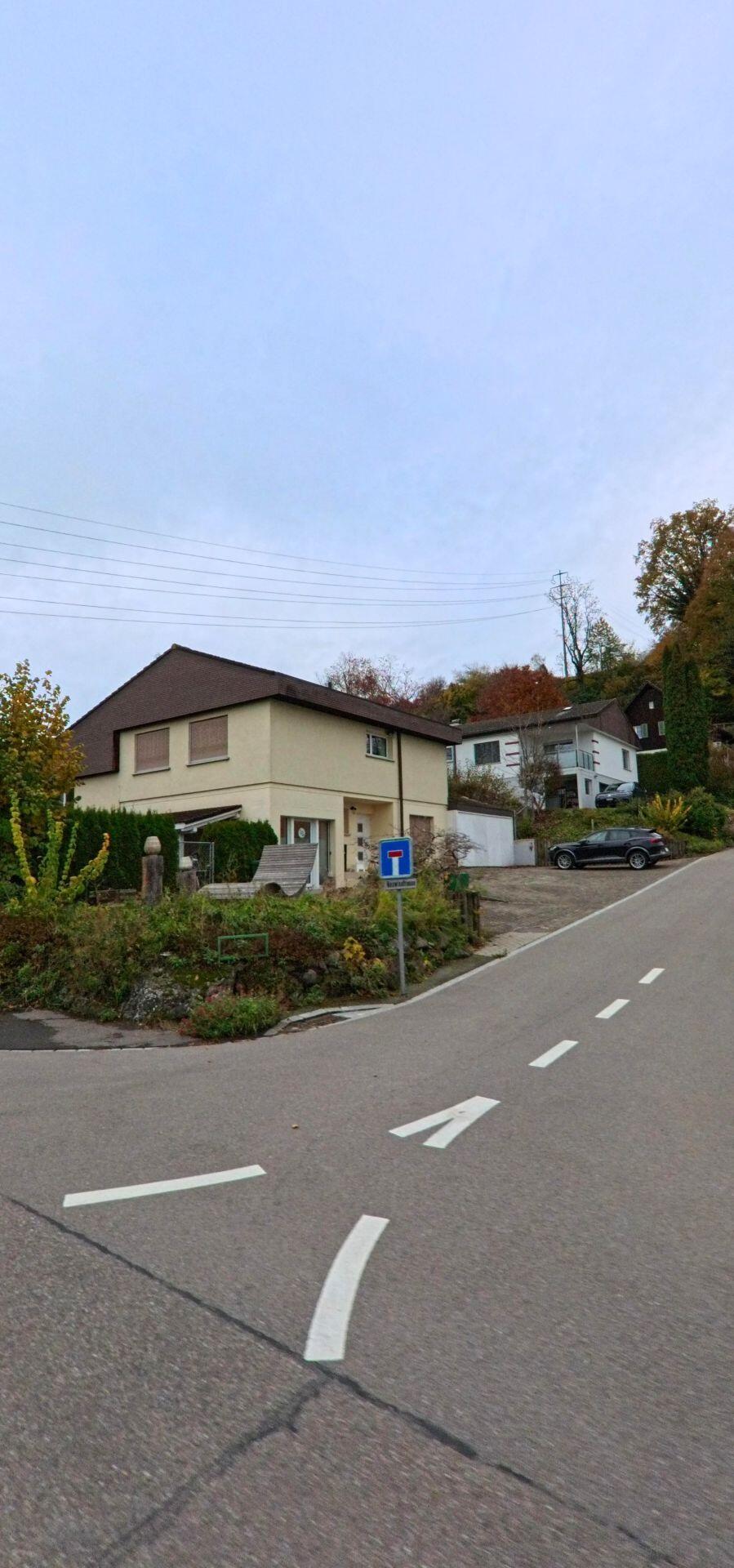Ein Wohnhaus am Hang in Kirchberg (9602, Kanton St. Gallen) steht an einer abzweigenden Strasse, waehrend weitere Hauser zwischen herbstlichen Baumen oberhalb liegen.