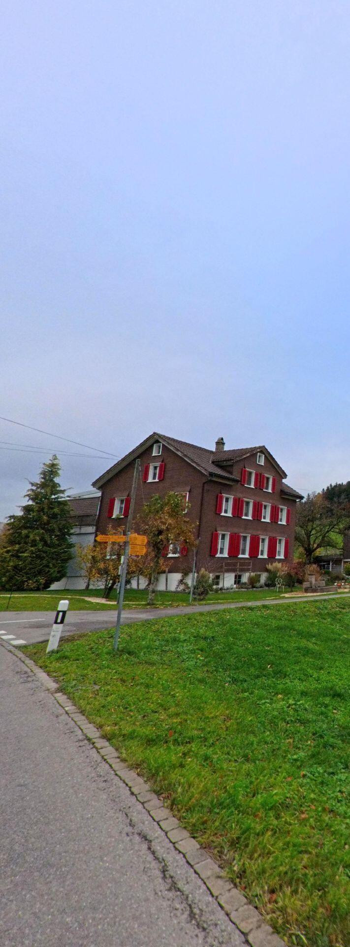 Ein braunes Wohnhaus mit roten Fensterlaeden steht in Kirchberg (9602, Kanton St. Gallen) neben einem Wegweiser an einer leicht geschwungenen Strasse mit angrenzender Gruenflaeche.