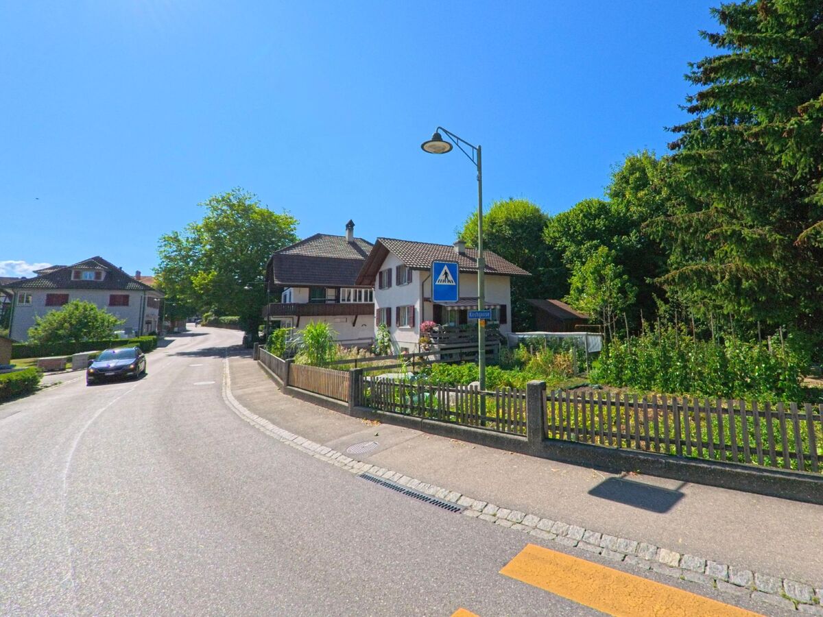 In Kirchdorf (3116, Kanton Bern) verläuft eine leicht ansteigende Strasse mit Fussgängerstreifen entlang von Einfamilienhäusern und Gärten, flankiert von hohen Bäumen am Waldrand.
