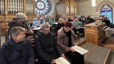 Mitglieder des Kirchenchors St. Mauritius Berikon (8965, Kanton Aargau) sitzen in einer Kirche mit Orgel und bunten Glasfenstern und bereiten sich auf den Gesang vor.