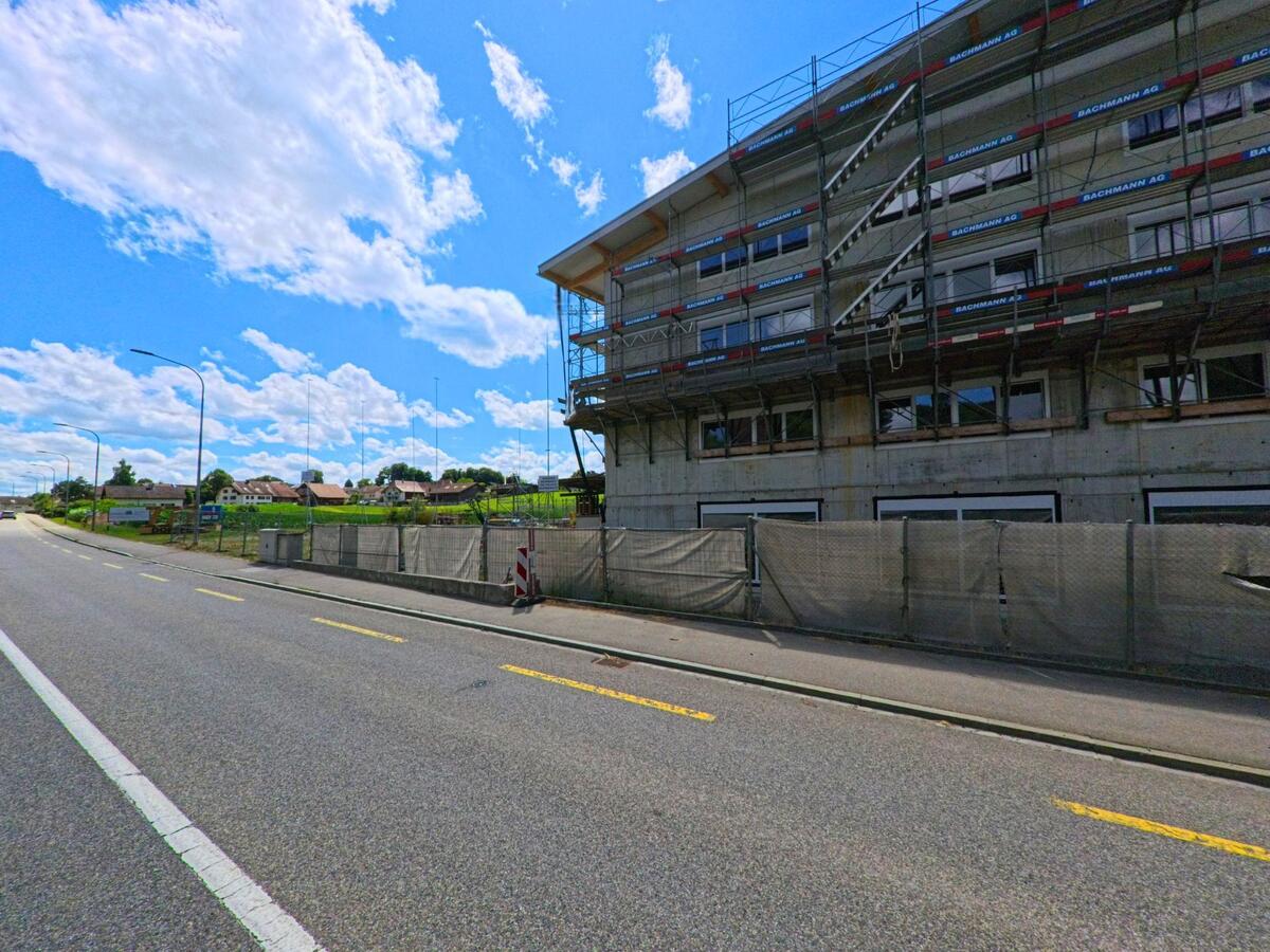 In Kirchleerau (5054, Kanton Aargau) entsteht an einer Hauptstrasse ein Neubau, der von Bauzaeunen und Geruesten umgeben ist, mit Blick auf ein Wohnquartier im Hintergrund.