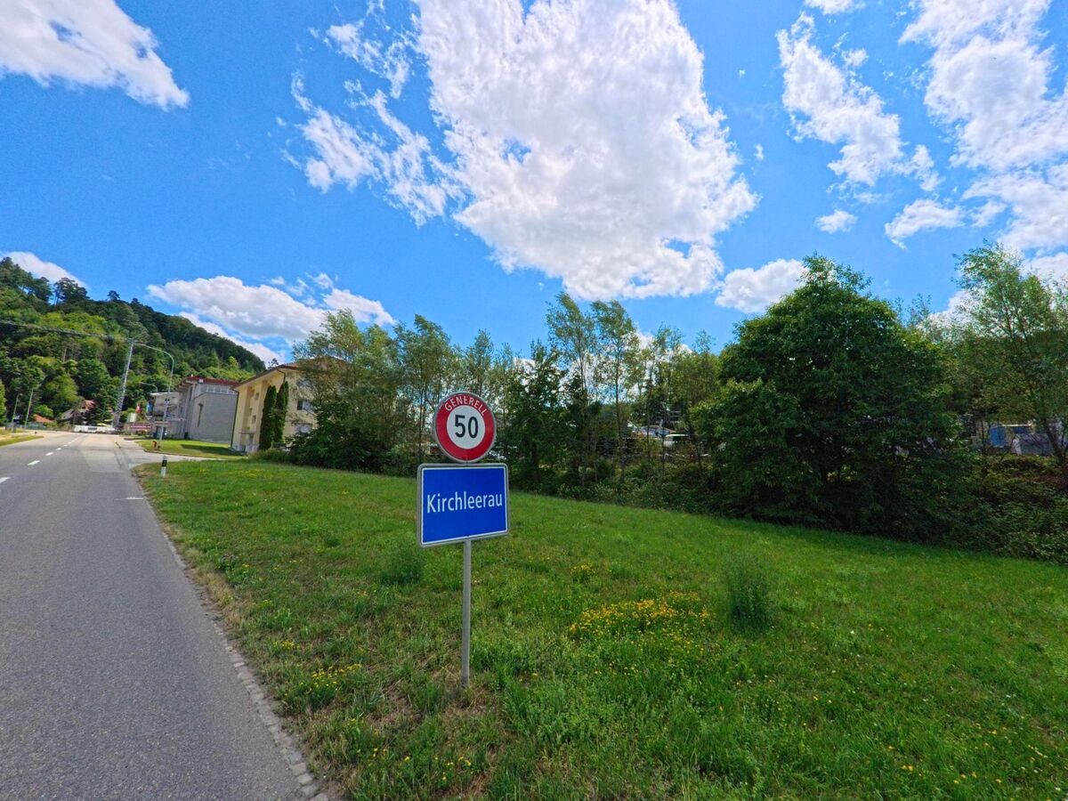 Am Ortseingang von Kirchleerau (5054, Kanton Aargau) steht ein blaues Ortsschild neben einer Landstrasse, flankiert von Wiese und Baeumen.