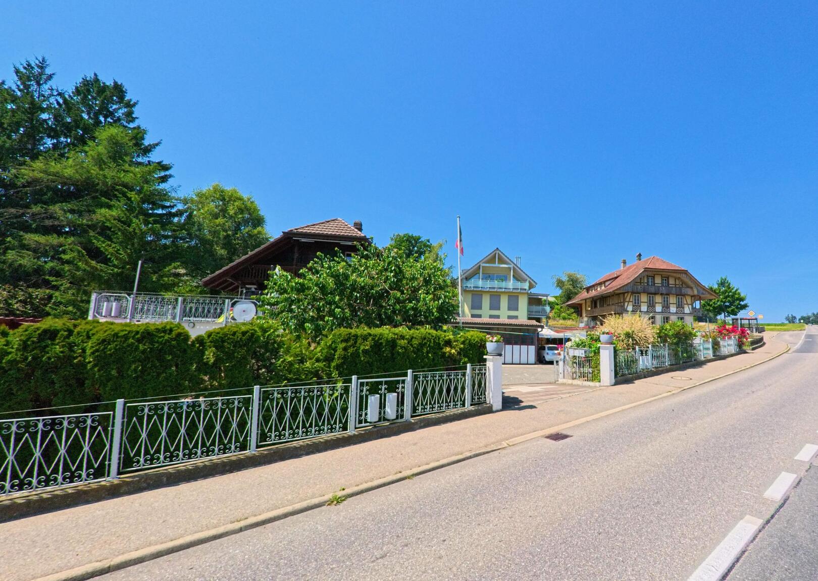 In Kirchlindach (3037, Kanton Bern) führt die Strasse an gepflegten Häusern mit Gärten und einem klaren, blauen Himmel vorbei, während die Strasse leicht ansteigt.