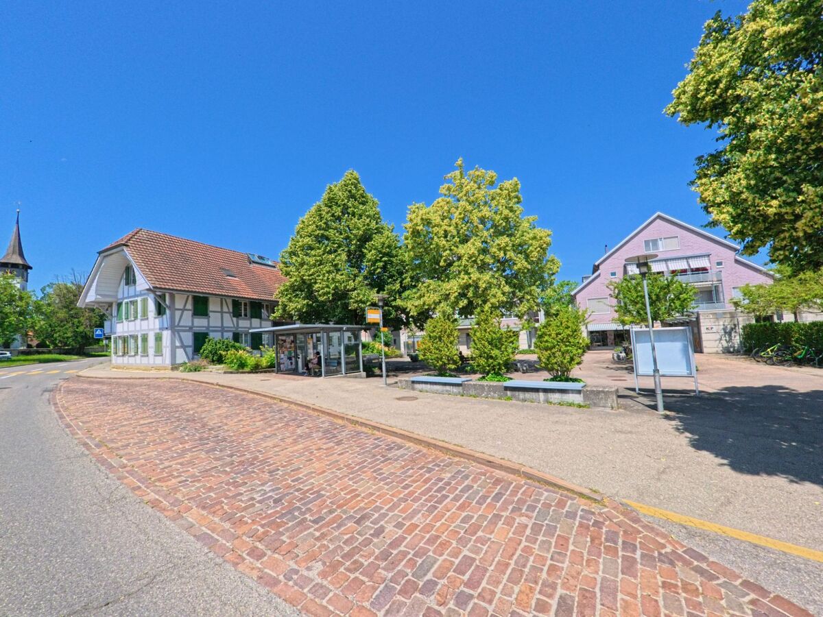 In Kirchlindach (3038, Kanton Bern) ist eine Straßenecke mit einem historischen Fachwerkhaus, einem modernen Gebäude und einer Bushaltestelle unter Bäumen abgebildet.