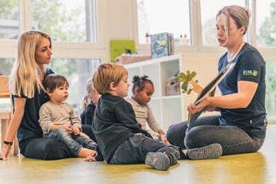 In Buchs (9470, Kanton St. Gallen) liest eine Betreuungsperson in einer Kita Kindern ein Bilderbuch vor, waehrend sie gemeinsam auf dem Boden sitzen.