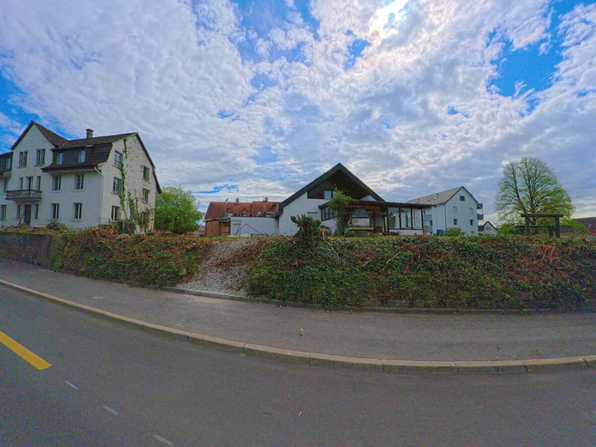 In Kleinandelfingen im Kanton Zürich (PLZ 8451) stehen mehrere Wohnhäuser inmitten einer leicht erhöhten Grünfläche, von der aus ein Schotterweg zur Strasse führt.