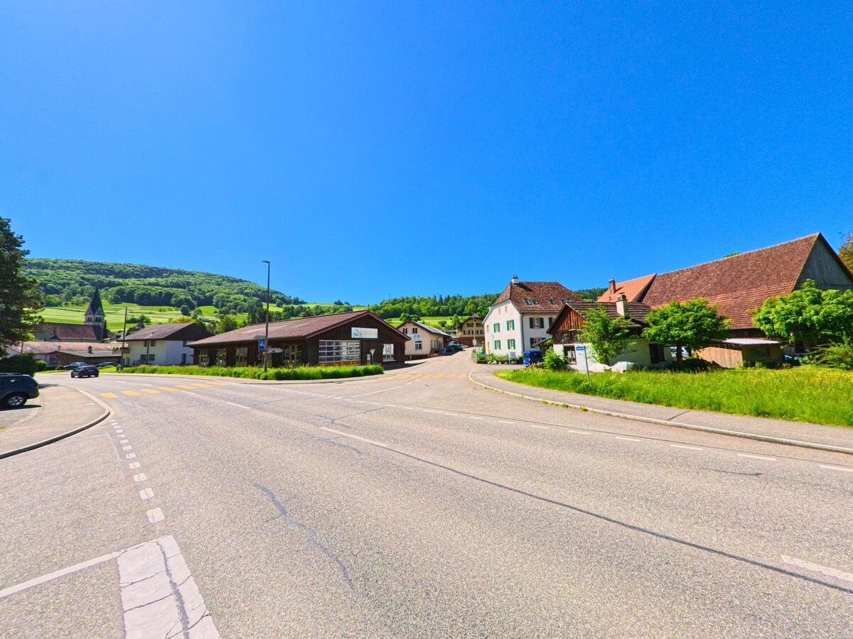 In Kleinlützel (4245, Kanton Solothurn) führt eine Straße an einem alten Gebäude und einer Kirche vorbei, mit grünen Hügeln im Hintergrund und strahlendem Sonnenschein.