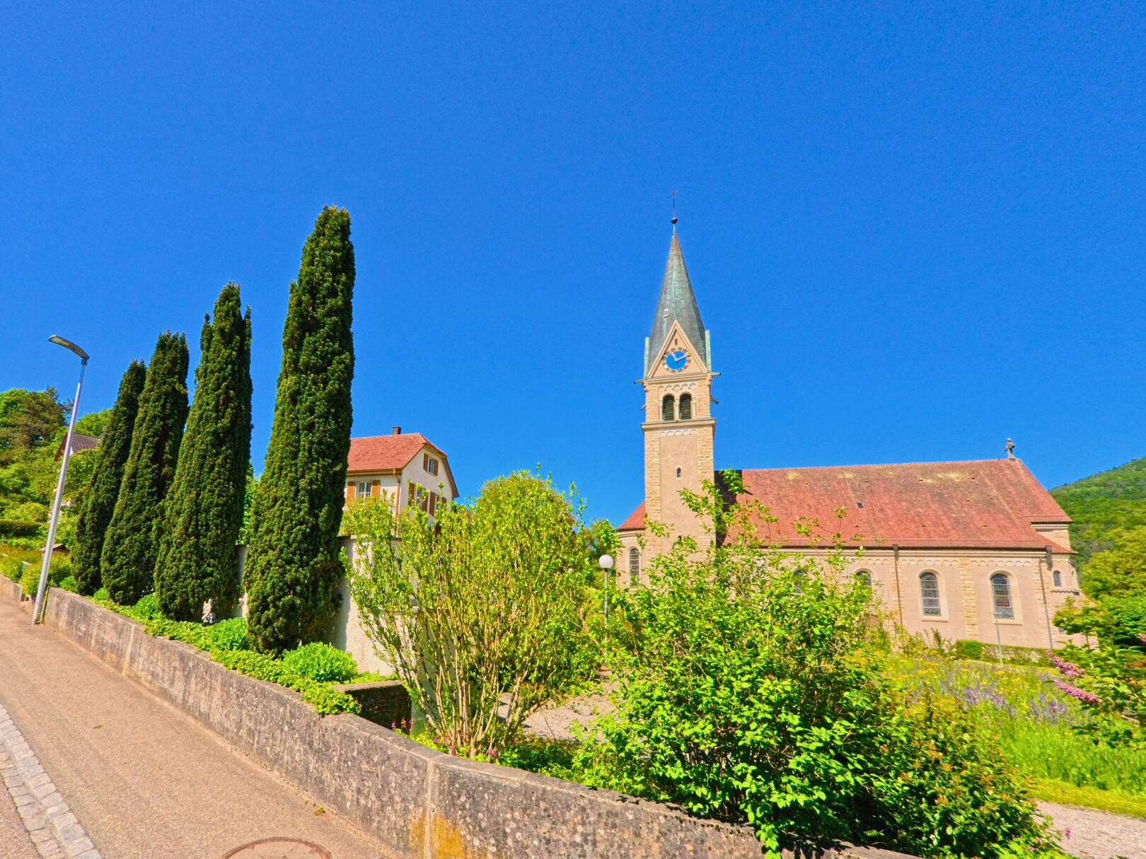 In Kleinlützel (4245, Kanton Solothurn) führt eine Strasse an einer Kirche mit einem markanten Turm und grünen Zypressen entlang, bei strahlendem Sonnenschein.