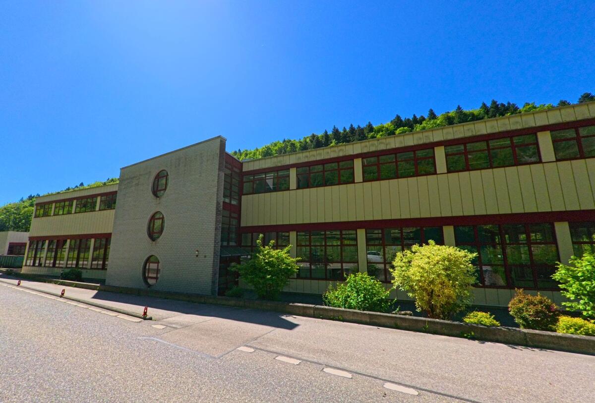 In Kleinlützel (4245, Kanton Solothurn) führt eine Straße an einem modernen Gebäude mit großen Fenstern und grünen Bäumen entlang, bei strahlendem Sonnenschein.