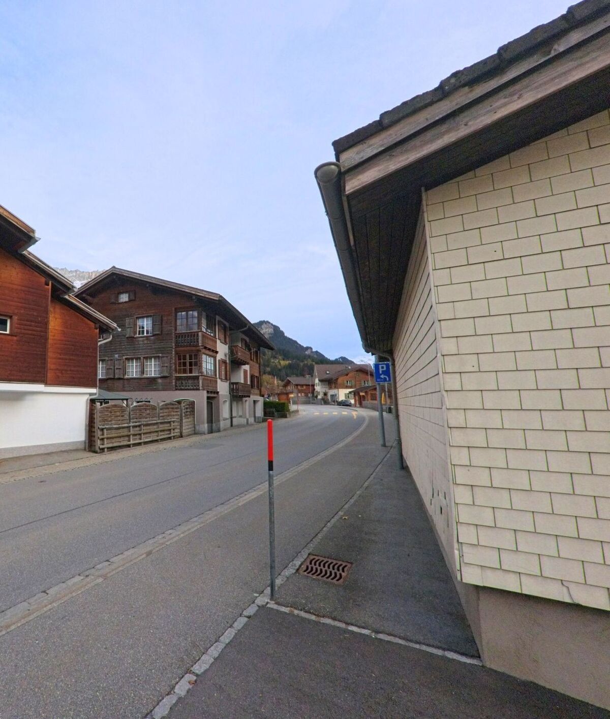 Entlang eines schmalen Trottoirs führt die Dorfstrasse an Holzhäusern vorbei durch Klosters (7247, Kanton Graubünden) mit Blick auf die umliegenden Berge.