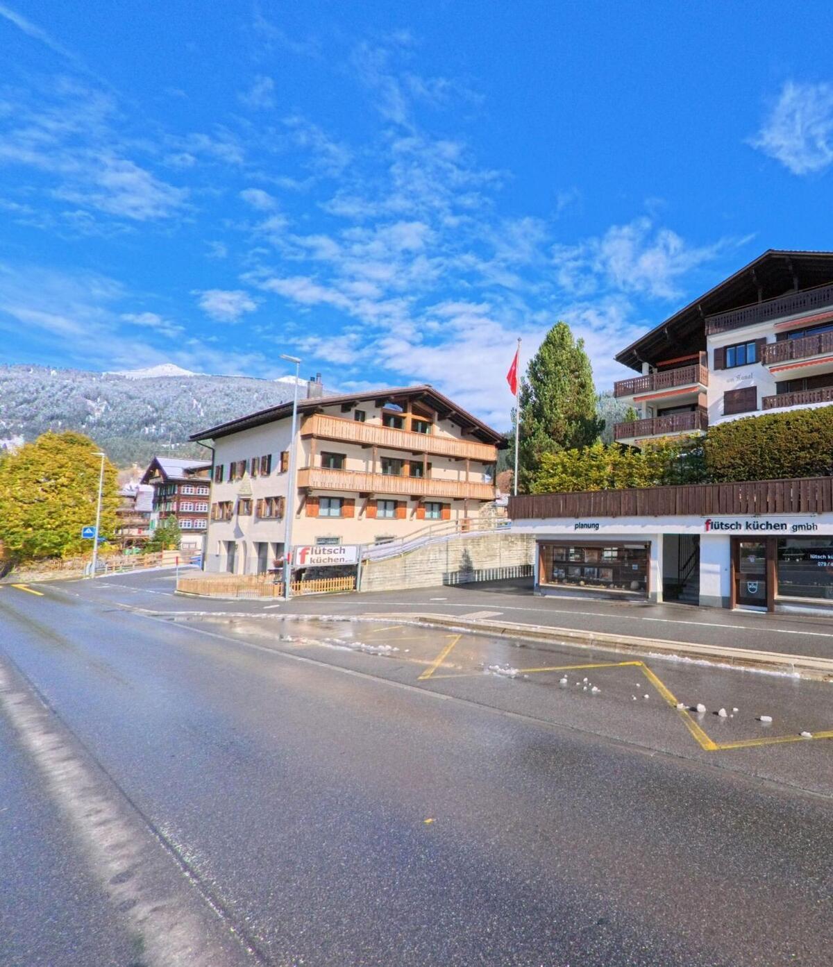 Entlang einer breiten Strasse mit Gewerbebauten und Wohnhäusern zeigt sich ein alltäglicher Dorfabschnitt von Klosters (7250, Kanton Graubünden) bei klarem Himmel.