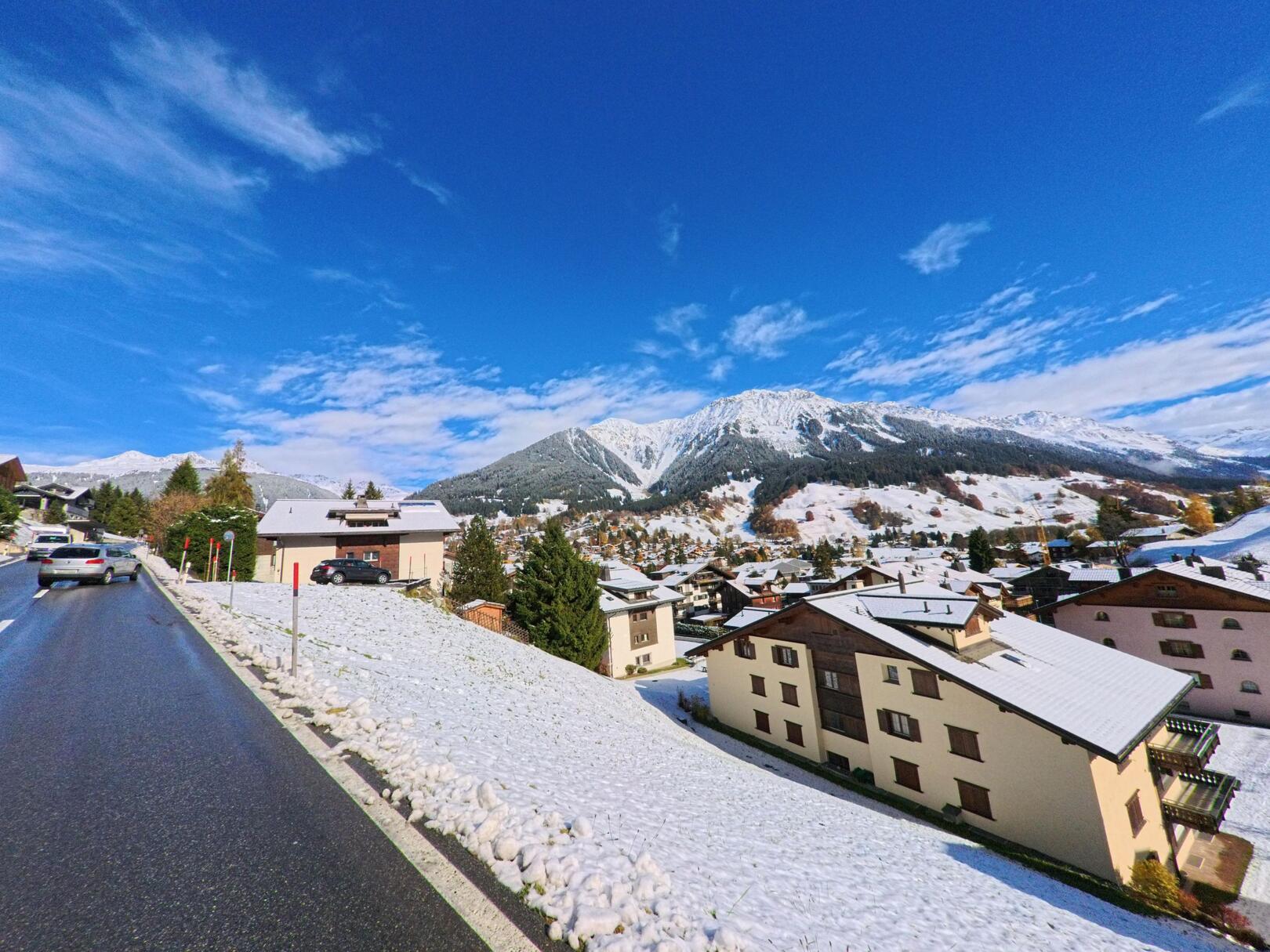 Von einer leicht ansteigenden Strasse aus zeigt sich das verschneite Siedlungsgebiet mit Bergpanorama in Klosters (7250, Kanton Graubünden) unter klarem Himmel.