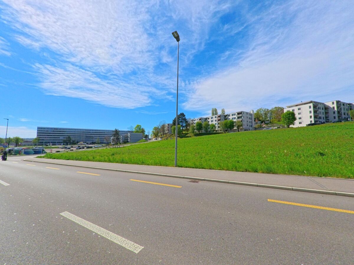 Eine breite Strasse in Kloten, PLZ 8302 im Kanton Zürich, verläuft an einer offenen Grünfläche mit dahinterliegenden Wohnsiedlungen und einem grösseren Verwaltungsgebäude.