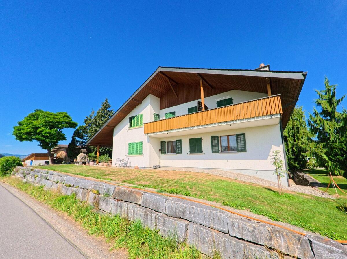 Am Ortsrand von Knutwil (6212, Kanton Luzern) steht ein Haus mit gruenen Fensterlaeden und grossem Balkon oberhalb einer Stuetzmauer aus Naturstein.