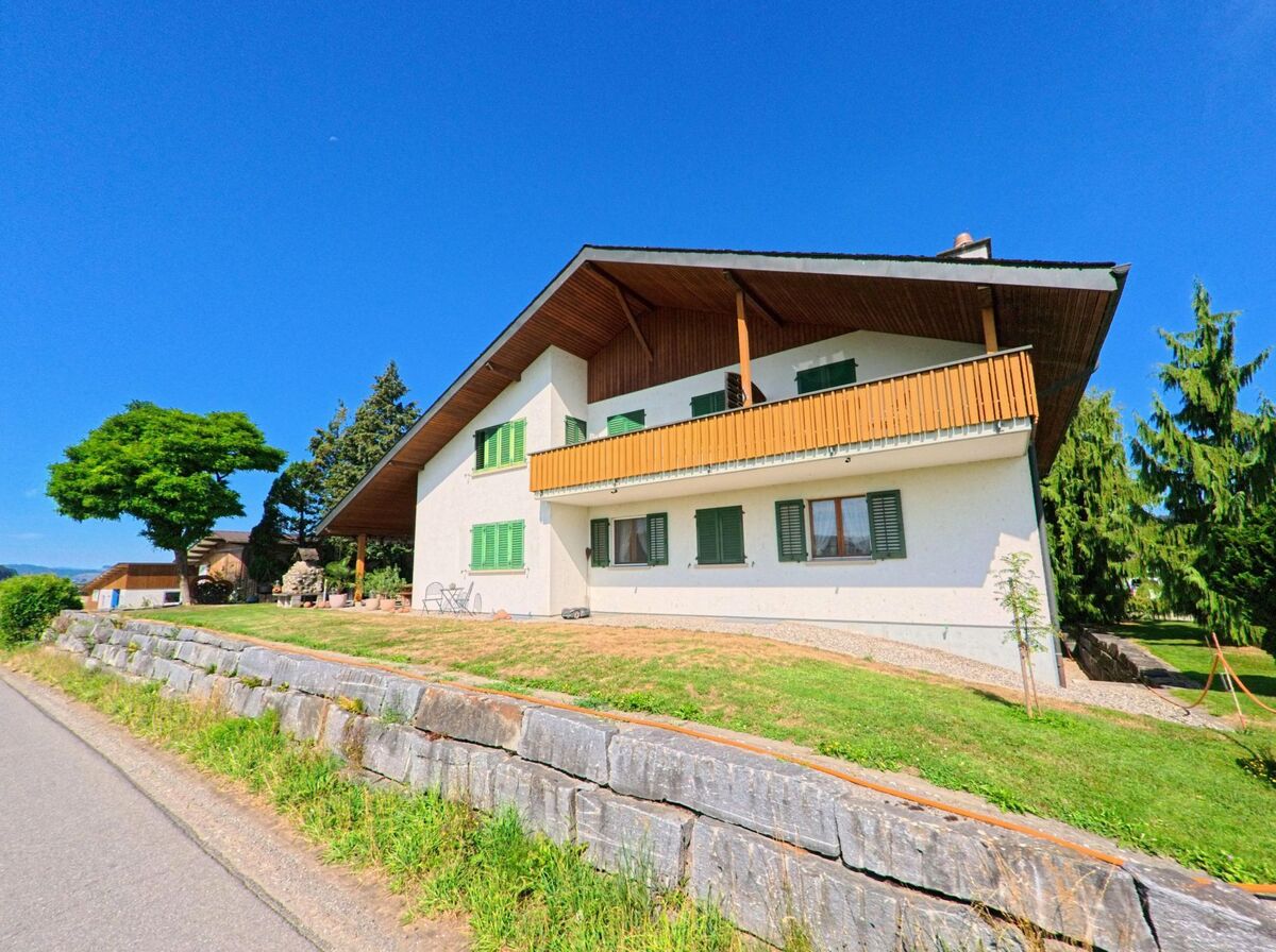Am Ortsrand von Knutwil (6212, Kanton Luzern) steht ein Haus mit gruenen Fensterlaeden und grossem Balkon oberhalb einer Stuetzmauer aus Naturstein.