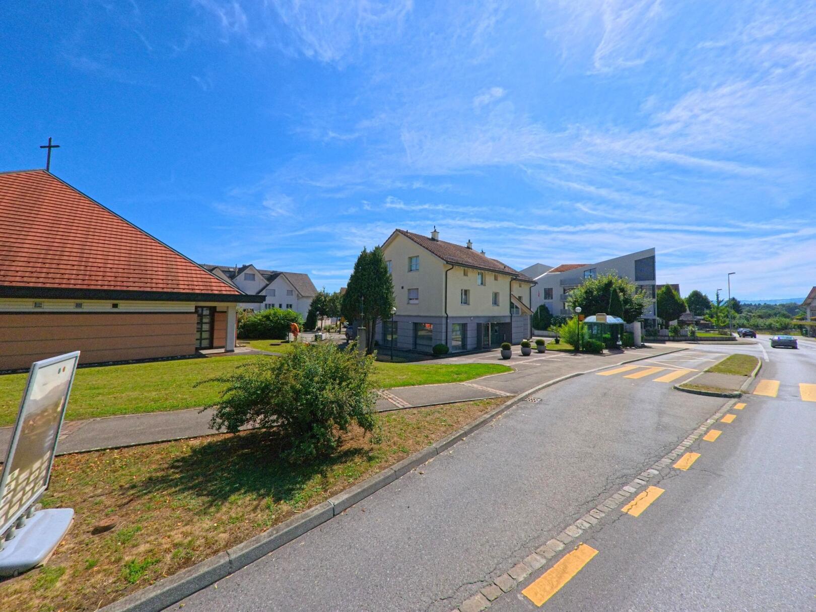 In Knutwil (6212, Kanton Luzern) liegt eine kleine Kirche mit rotem Ziegeldach direkt an einer Strasse mit Fussgaengerstreifen und moderner Wohnbebauung im Hintergrund.