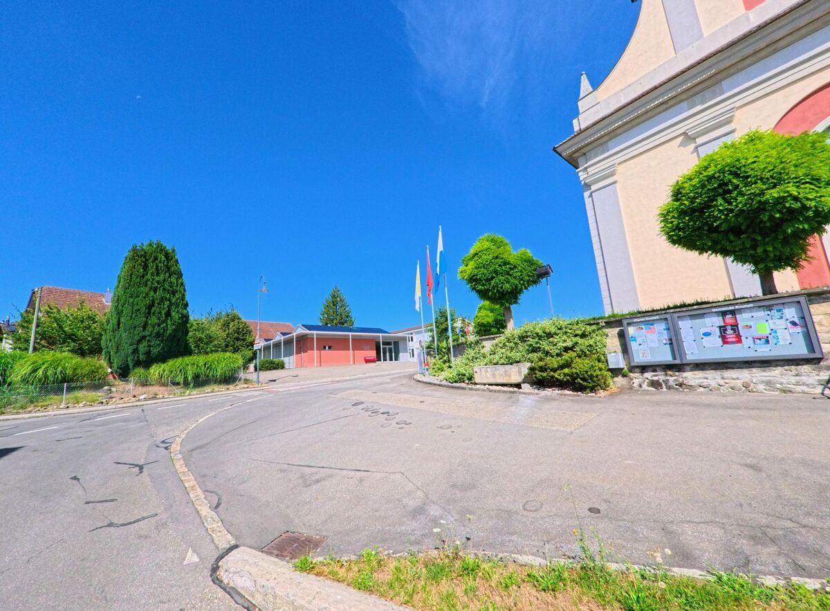 In Knutwil (6213, Kanton Luzern) liegt eine Kirche mit gepflegter Umgebung an einer leicht ansteigenden Strasse, neben der ein Schaukasten mit Gemeindeinformationen steht.