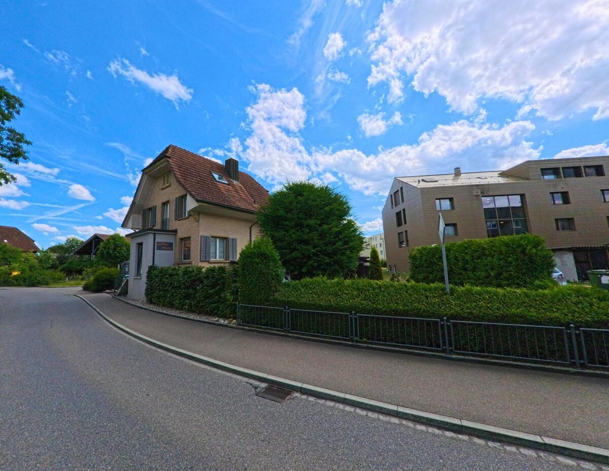 In einer Wohnstrasse von Kölliken (5742, Kanton Aargau) stehen ein älteres Einfamilienhaus mit rotem Ziegeldach und ein moderner Wohnblock in direkter Nachbarschaft.