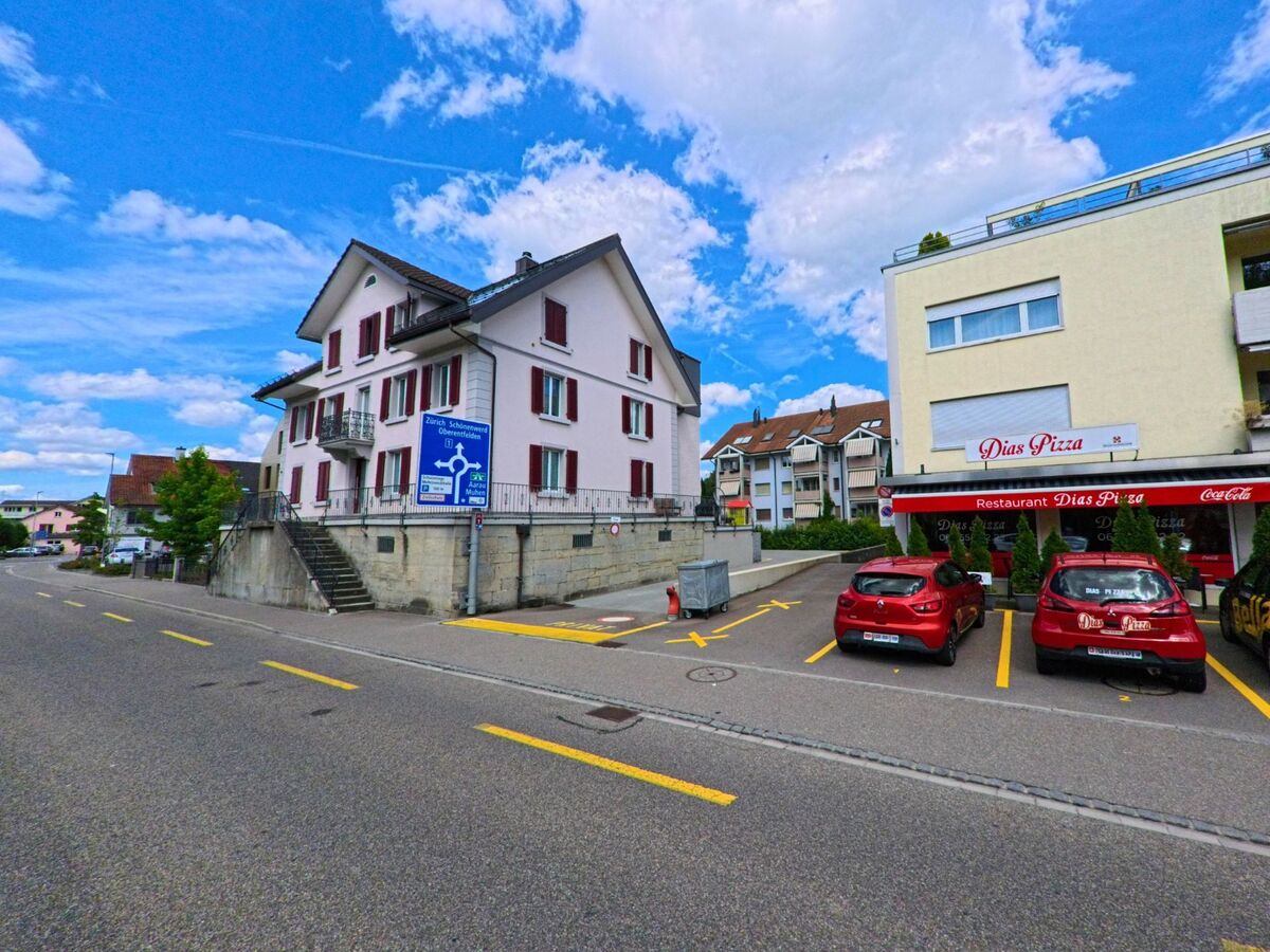 In Kölliken (5742, Kanton Aargau) steht an einer Hauptstrasse ein Wohnhaus mit Wegweiser und daneben das Restaurant „Dias Pizza“ mit Parkplätzen und markanten roten Autos.