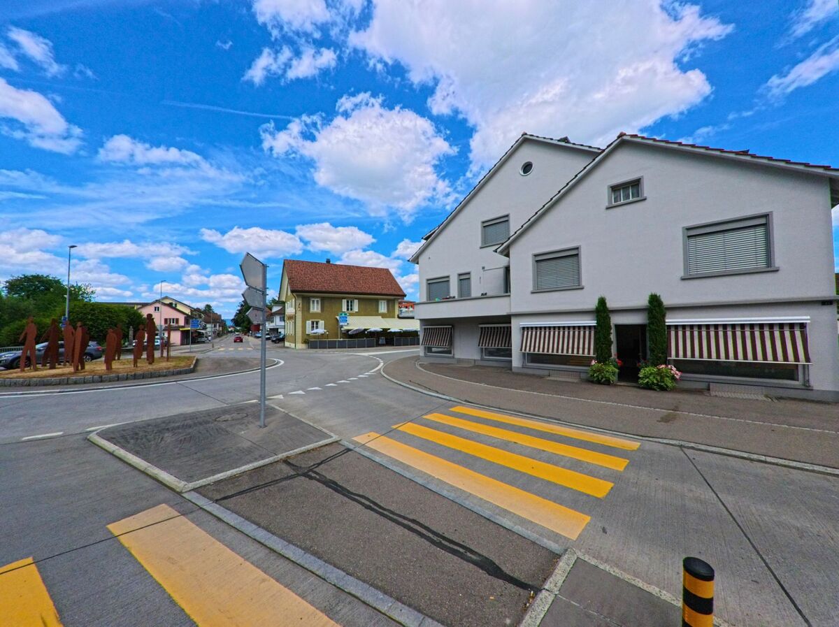 In Kölliken (5742, Kanton Aargau) verläuft eine Strasse mit Fussgängerstreifen entlang mehrerer Wohnhäuser, während auf einer Verkehrsinsel rostrote Figurenskulpturen stehen.