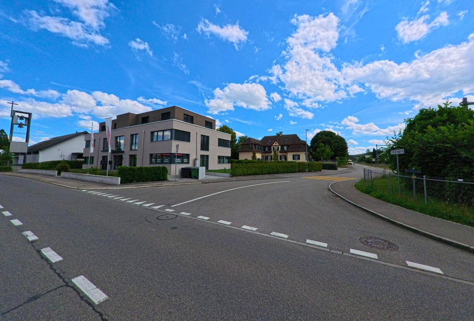 An einer Strassenkreuzung in Kölliken (5742, Kanton Aargau) stehen ein modernes Wohn- und Geschäftshaus sowie ein traditionelles Wohnhaus unter leicht bewölktem Himmel.
