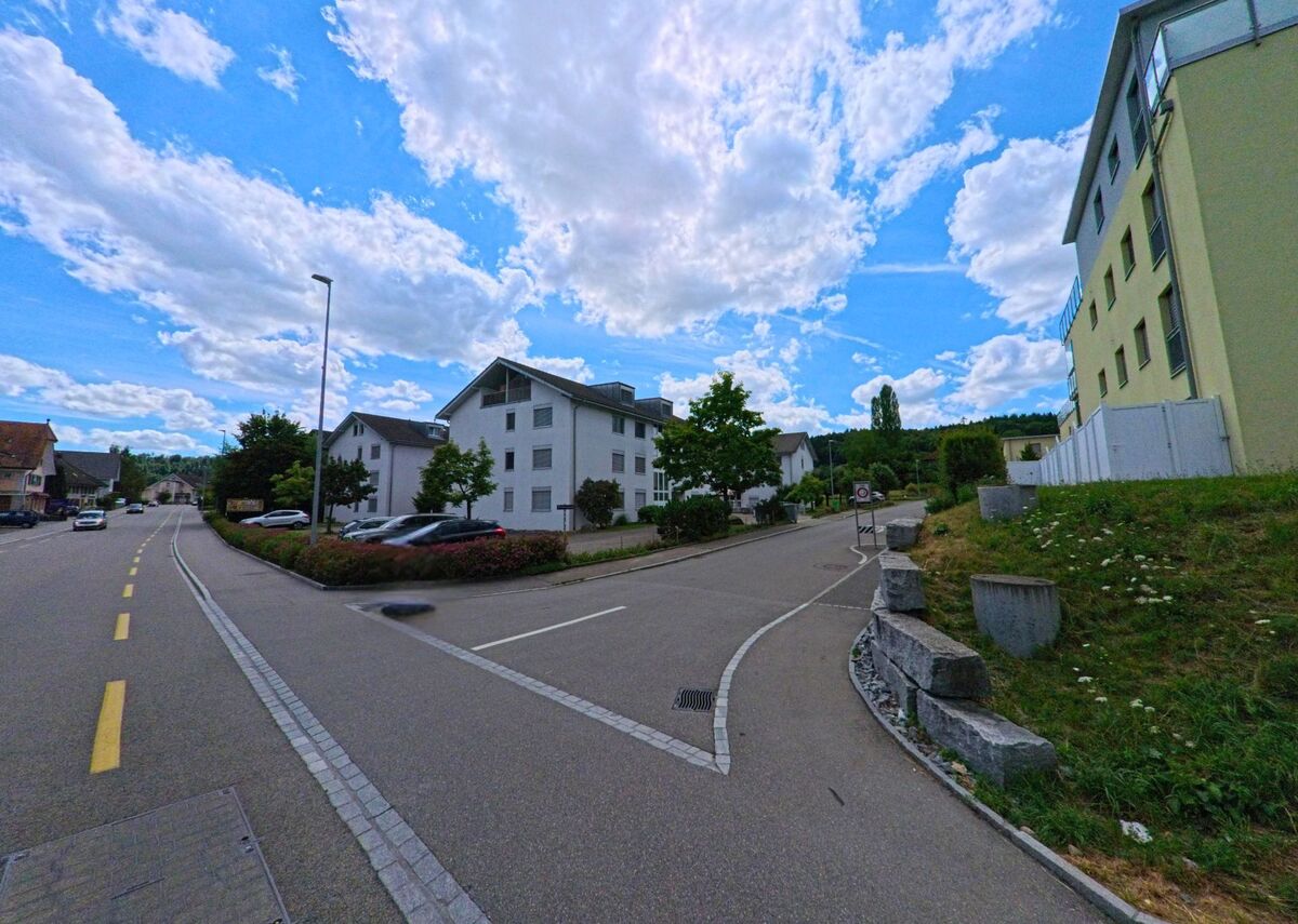 In einem Wohngebiet von Kölliken (5742, Kanton Aargau) verlaufen zwei Strassen zwischen modernen Mehrfamilienhäusern, flankiert von Grünflächen und bepflanzten Rabatten.