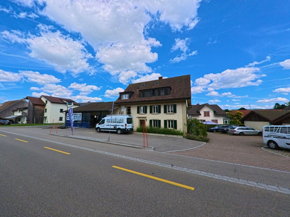 An einer Durchgangsstrasse in Kölliken (5742, Kanton Aargau) befinden sich ein Wohnhaus und ein Werkstattgebäude mit der Aufschrift „mön metallbau ag“, umgeben von parkierten Fahrzeugen.