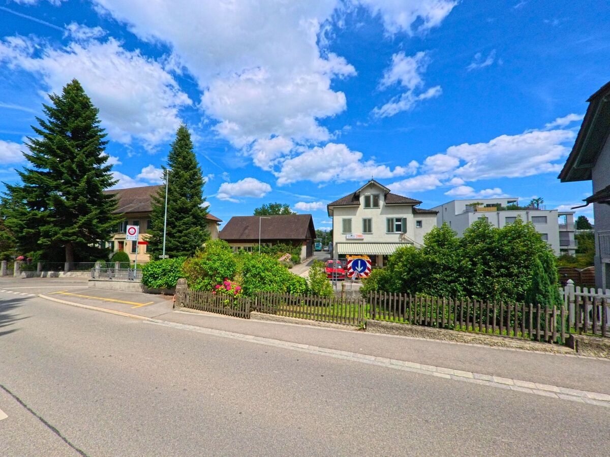 In einem Wohnquartier von Kölliken (5742, Kanton Aargau) stehen zwischen gepflegten Gärten und Tannenbäumen mehrere Ein- und Mehrfamilienhäuser unter blauem Himmel.