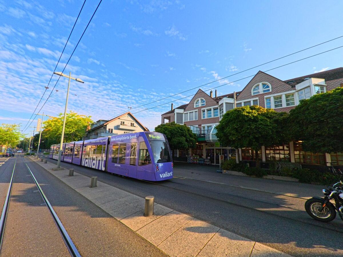 Eine lilafarbene Strassenbahn faehrt durch Koeniz (3084, Kanton Bern) an einer von Baeumen gesaeumten Strasse mit Geschaeften in traditionellen Gebaeuden vorbei.