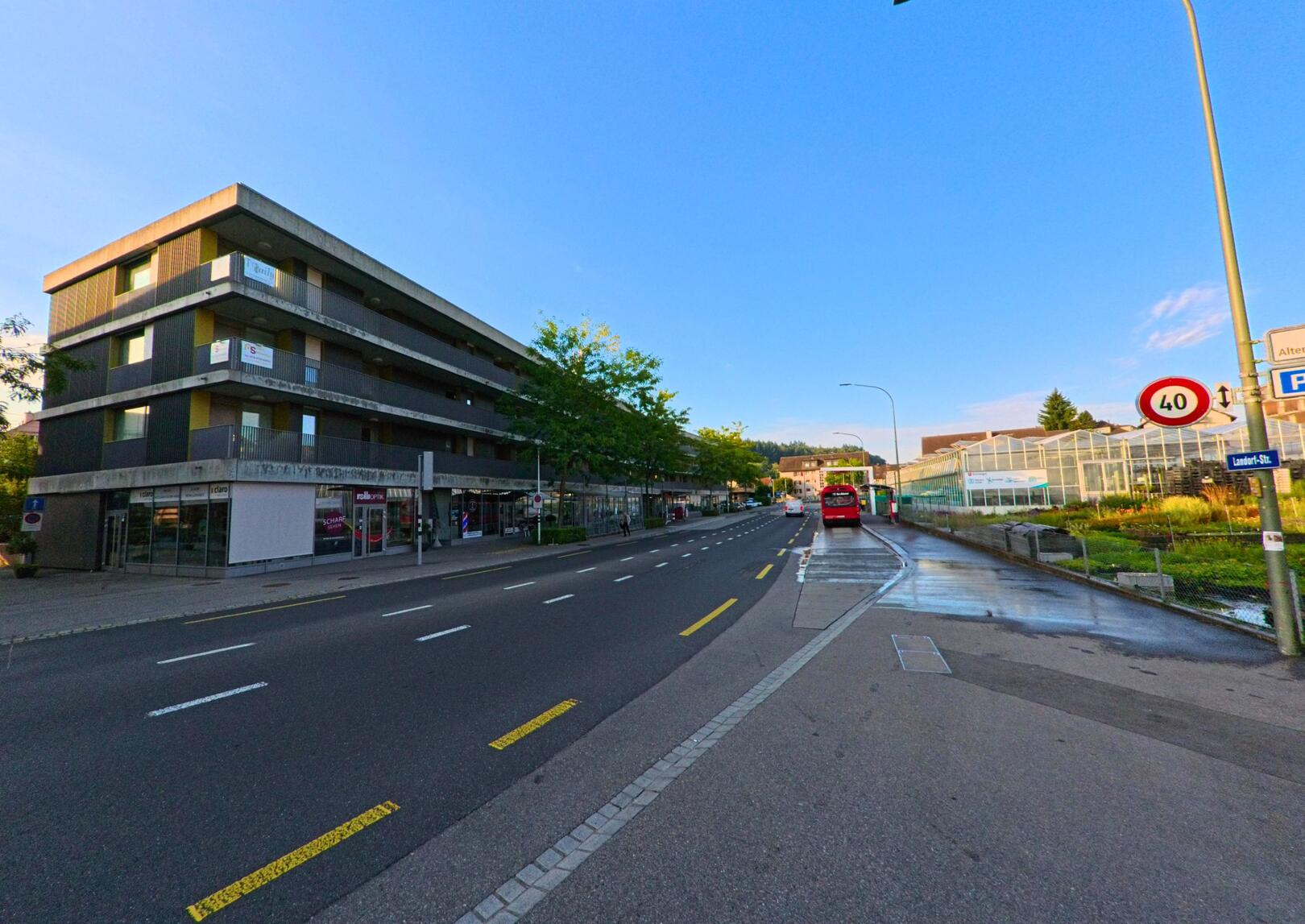 Eine breite Strasse in Koeniz (3098, Kanton Bern) verlaeuft zwischen einem modernen Wohn- und Geschaeftsgebaeude und einem Gewaechshaus, waehrend ein roter Bus in der Ferne haelt.