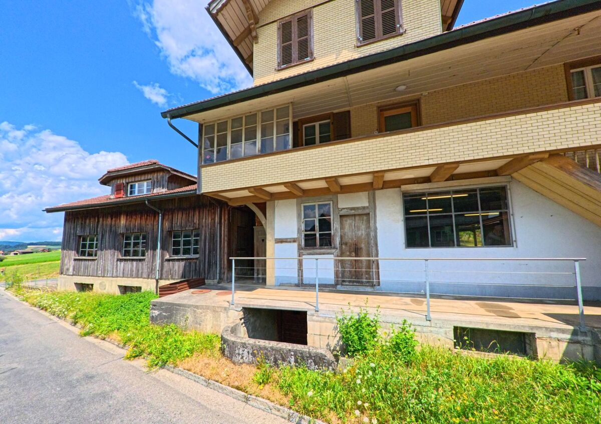 In Konolfingen (PLZ 3503, Kanton Bern) steht ein Gebäude mit gemischter Holz- und Backsteinfassade an einer schmalen Strasse mit Blick auf die offene Landschaft.