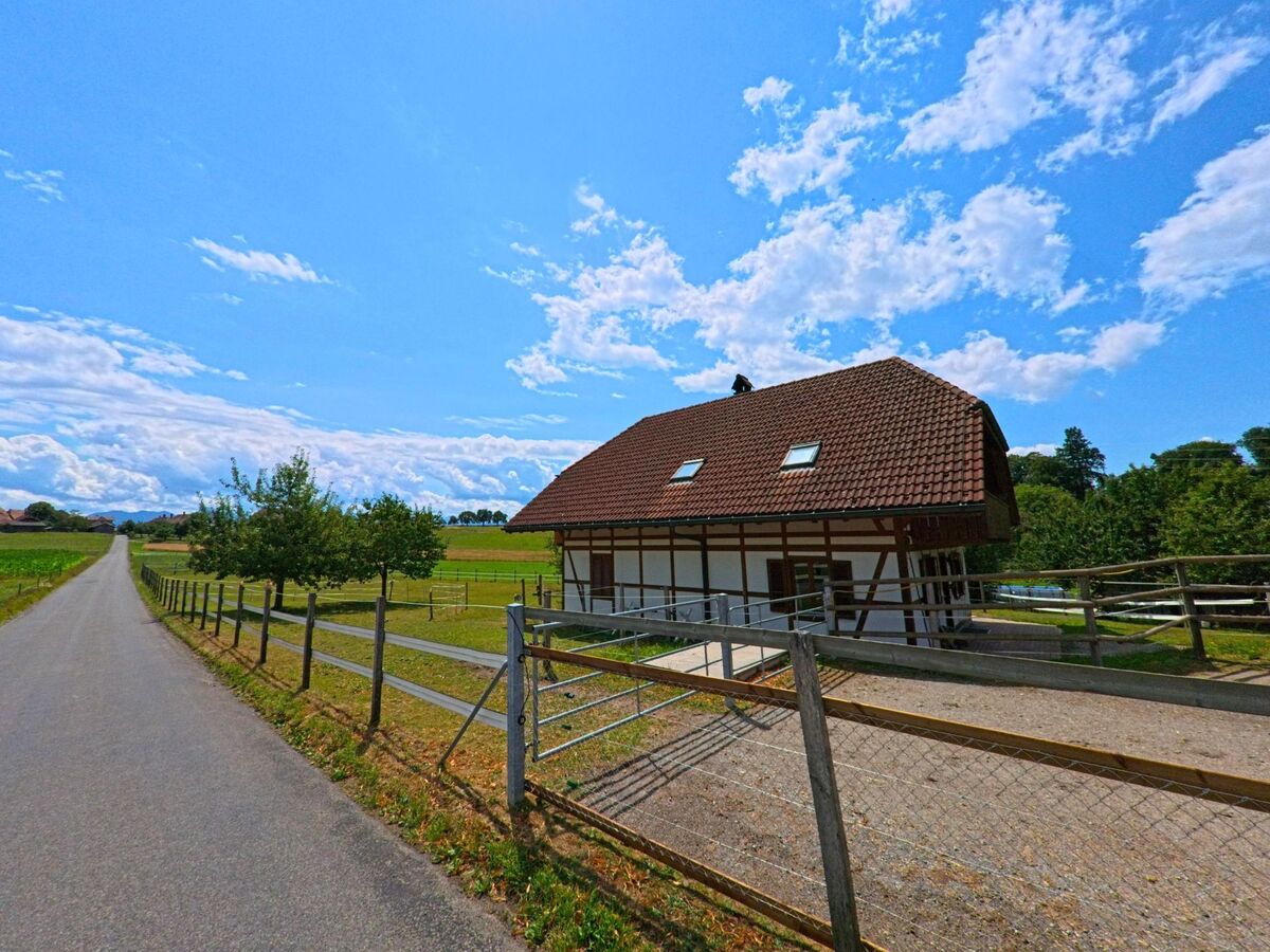 Am Rand von Konolfingen (3510, Kanton Bern) führt ein schmaler Asphaltweg an einem umzäunten Fachwerkhaus mit rotem Ziegeldach vorbei, eingebettet in eine offene Feldlandschaft.