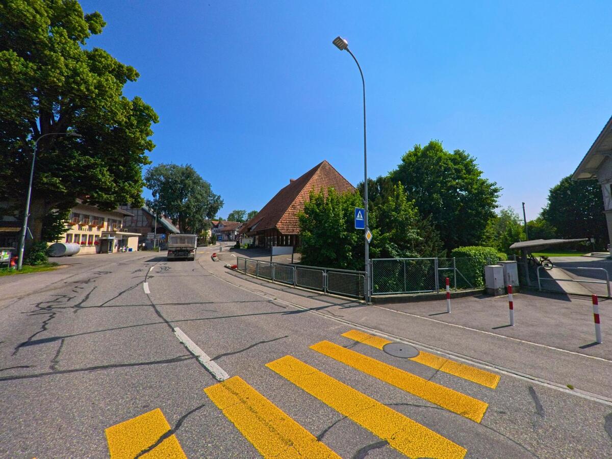 In Koppigen (3425, Kanton Bern) verläuft die Hauptstrasse mit einem markanten Zebrastreifen entlang eines alten Gebäudes mit steilem Walmdach und schattenspendenden Bäumen.