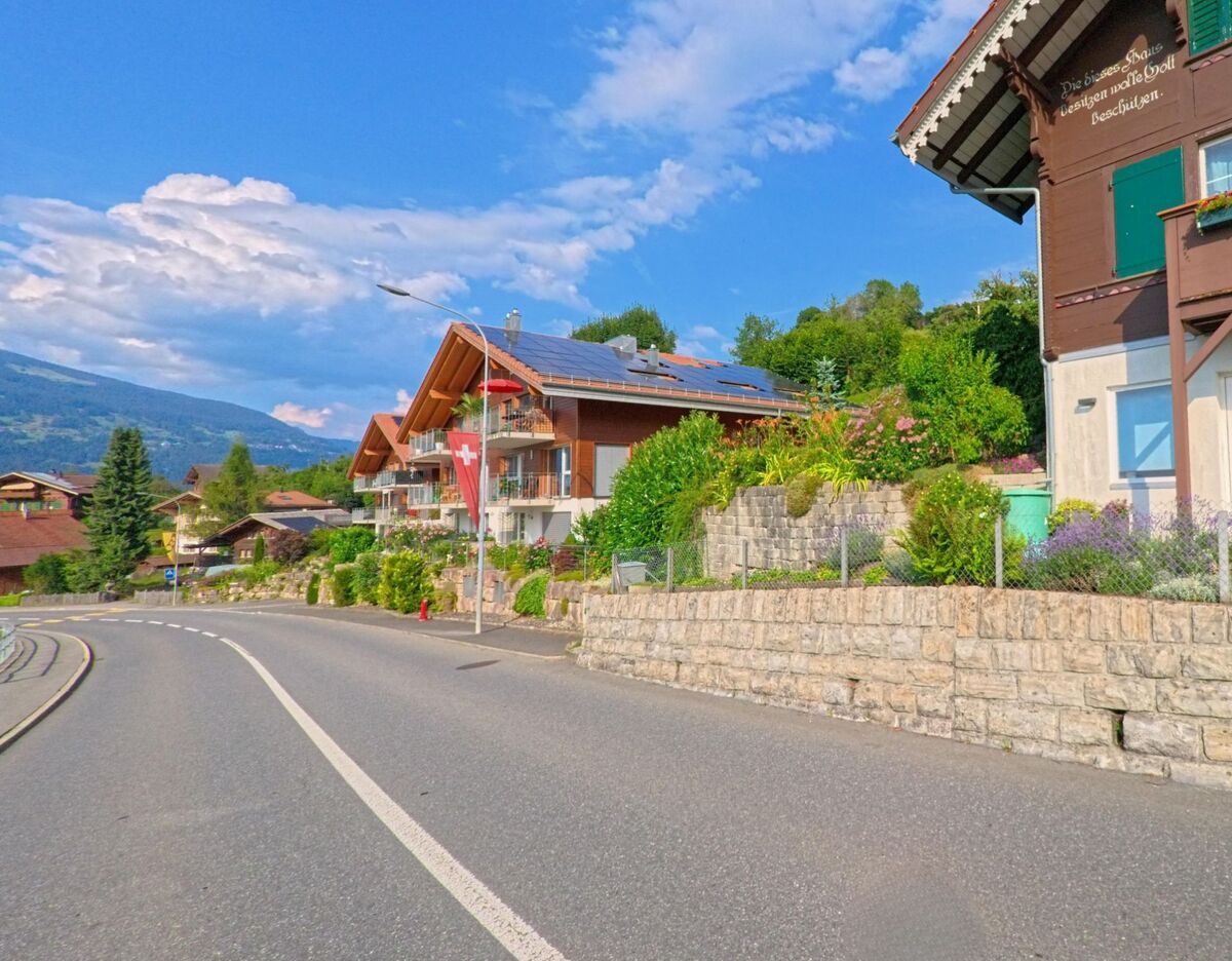 In Krattigen (3704, Kanton Bern) säumen gepflegte Mehrfamilienhäuser mit Solaranlagen und Vorgärten die leicht geschwungene Strasse, während eine Schweizer Fahne im Wind weht.