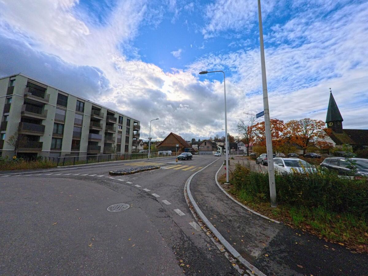 Auf einer breiten Strasse in Kreuzlingen (8280, Kanton Thurgau) führt ein gelber Fussgängerübergang an einer Verkehrsinsel vorbei, während im Hintergrund Wohnblocks und ein Kirchturm zu sehen sind.