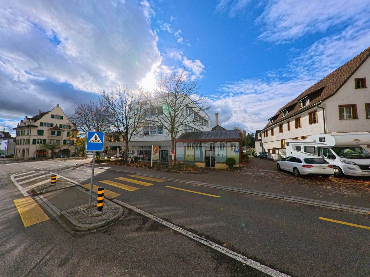 Bei einem Fussgängerübergang in Kreuzlingen (8280, Kanton Thurgau) steht ein blaues Zebrastreifen-Schild vor einer Kreuzung mit Gebäuden und parkenden Fahrzeugen.