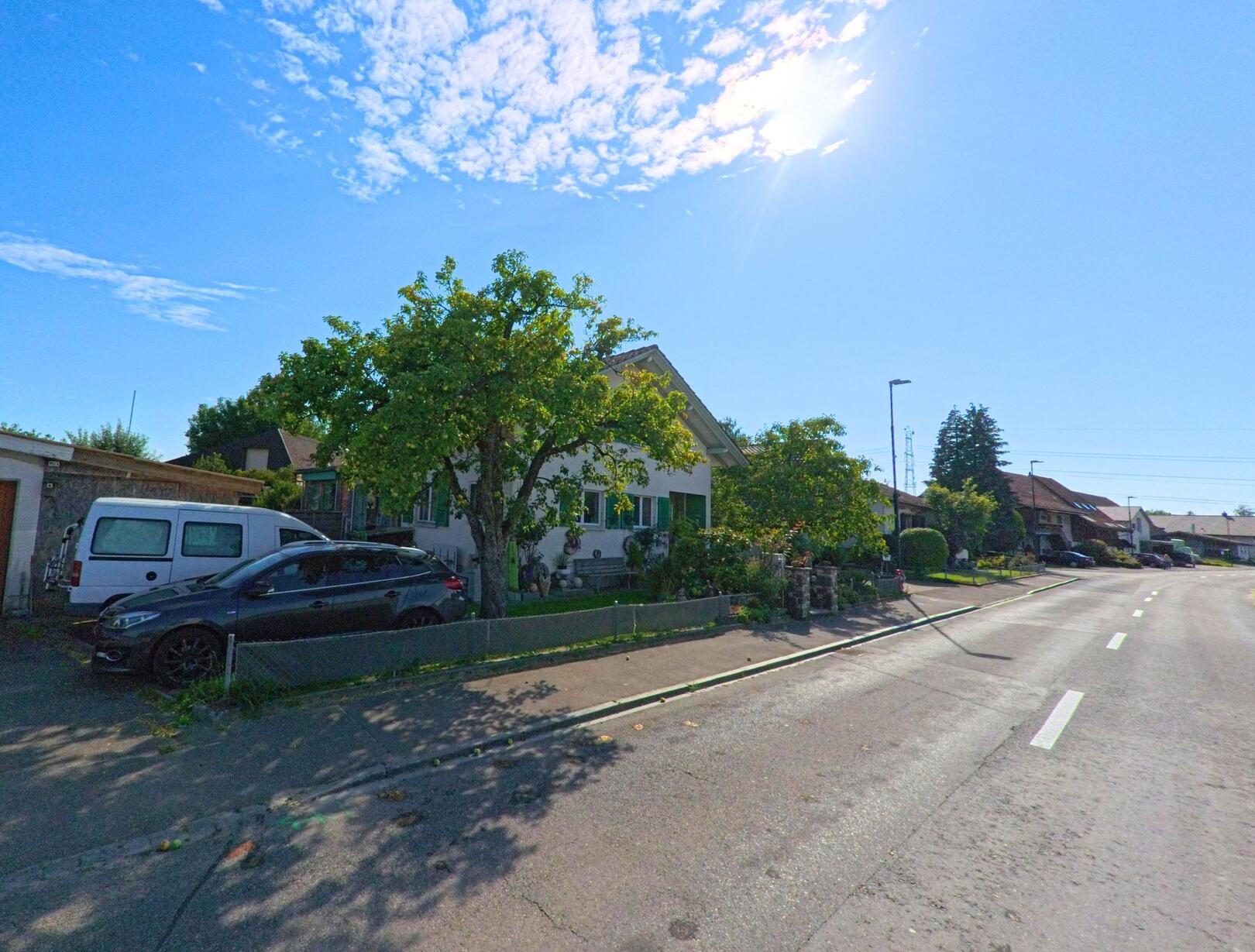 An einer breiten Strasse in Kriechenwil (3179, Kanton Bern) steht ein Einfamilienhaus mit Vorgarten und geparkten Fahrzeugen in der Einfahrt.