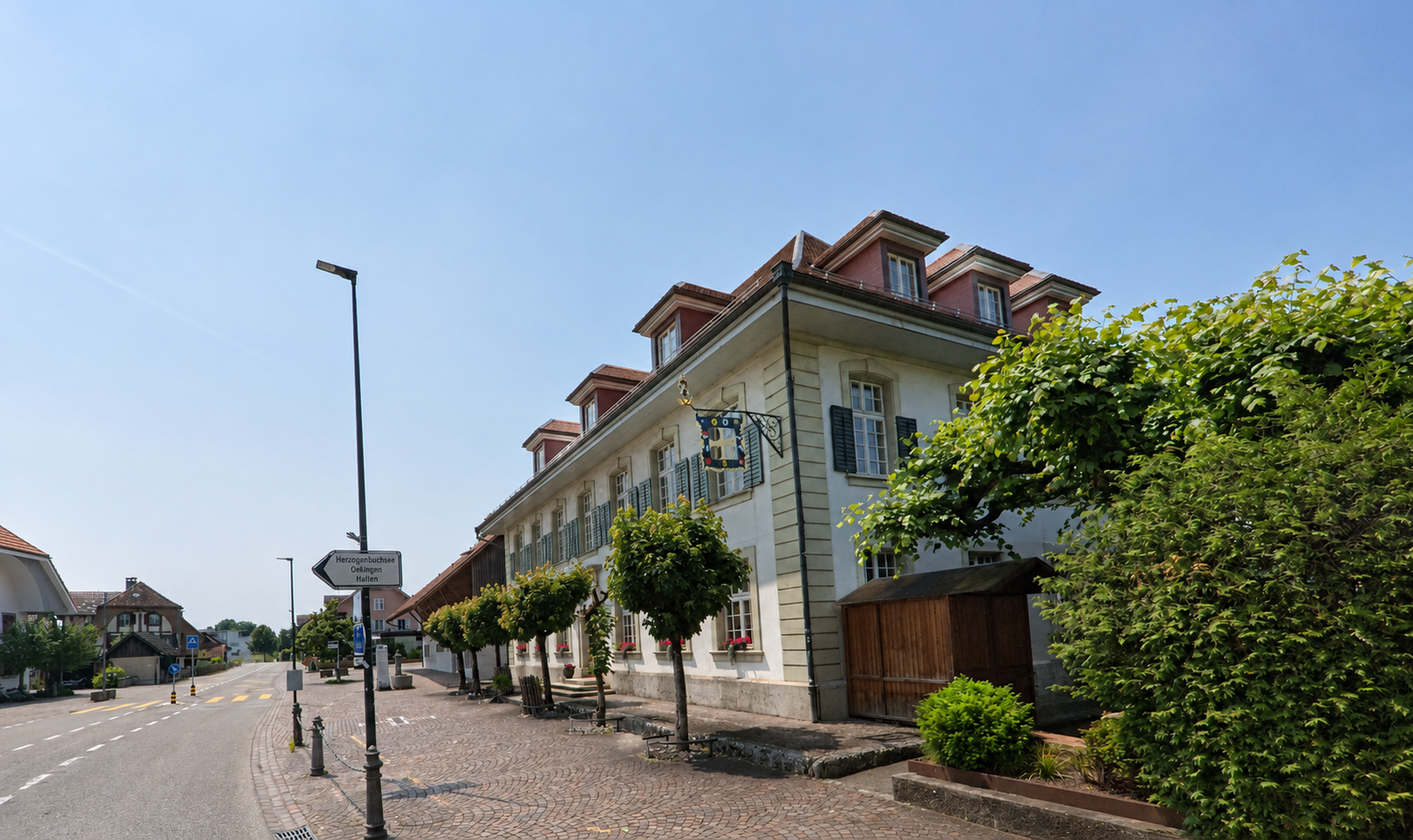 In Kriegstetten (4566, Kanton Solothurn) führt eine gepflasterte Strasse an einem historischen Gebäude mit bunten Fensterläden und grünen Bäumen entlang.