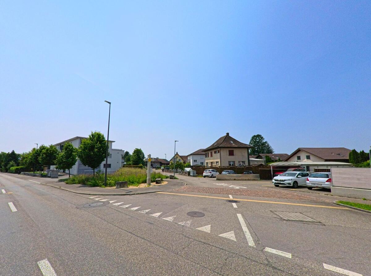 Eine Straße in Kriegstetten (4566, Kanton Solothurn) führt an modernen Häusern entlang, mit einer Straßenecke, die von Bäumen gesäumt ist und einer Einfahrt auf der rechten Seite.
