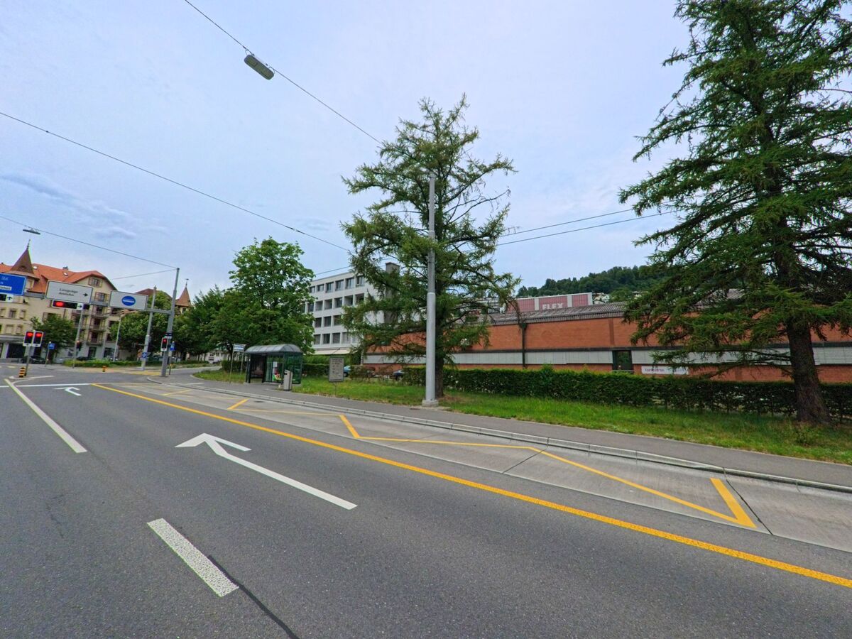 In Kriens (6010, Kanton Luzern) zeigt die Strasse eine Bushaltestelle sowie Baeume und Gebaeude entlang der breiten Fahrbahn, die von Verkehrsschildern und markierten Fahrspuren begleitet wird.