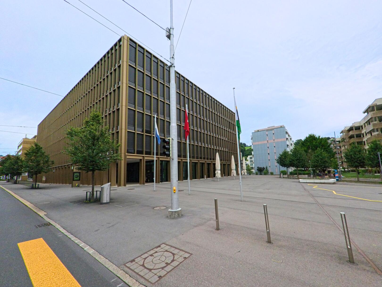 In Kriens (6010, Kanton Luzern) steht ein modernes Verwaltungsgebäude mit markanten Fassaden und Fahnen vor einer weiten, offenen Platzfläche, umgeben von Bäumen und modernen Wohngebäuden.