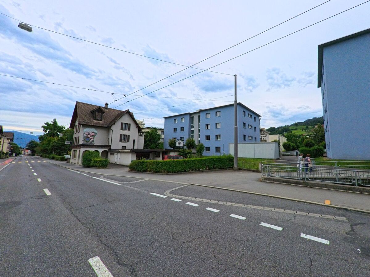 In Kriens (6010, Kanton Luzern) stehen zwei moderne Wohnblocks neben einem traditionellen Haus, während Spaziergänger die Strasse entlanggehen, gesäumt von gepflegten Grünflächen.