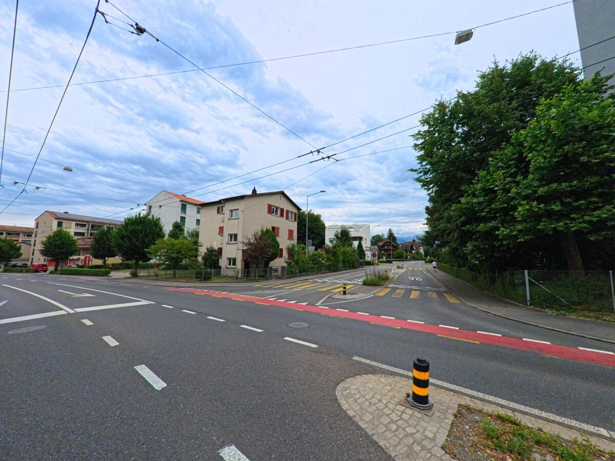 In Kriens (6010, Kanton Luzern) zeigt eine ruhige Strasse, gesäumt von Wohnhäusern und Bäumen, einen Zebrastreifen und eine Verkehrskontrolle, während der Blick in die Ferne von den Alpen geprägt ist.